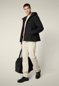 Hombre con chaqueta negra, pantalones beige y botas negras, sosteniendo un bolso de deporte negro, de pie contra un fondo neutro.