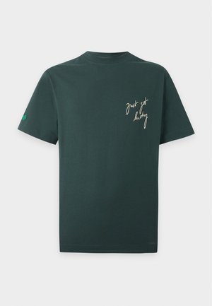 Homme à la tête rasée portant un t-shirt vert foncé avec un texte blanc "just get lucky,", jean bleu, ceinture noire et baskets blanches, debout avec une main dans la poche.