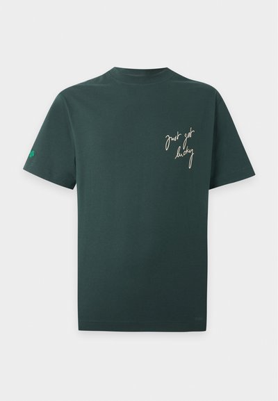 T-shirt manches courtes vert foncé avec texte manuscrit couleur crème "just got lucky" sur la poitrine gauche et un petit trèfle à quatre feuilles vert sur la manche gauche.