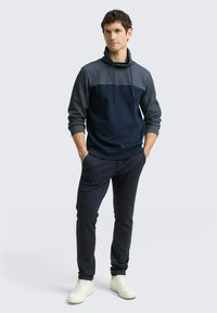 Niebiesko-szara bluzka z wysokim kołnierzem i sznurkiem, zestawiona z ciemnymi spodniami i białymi trampkami. Gładka faktura tkaniny.