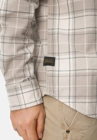 Beige Flanellhemd mit blauem Karo-Muster, ausgestattet mit geknöpften Manschetten und einem Etikett am Saum mit der Aufschrift "MADE FOR DAILY ADVENTURES".