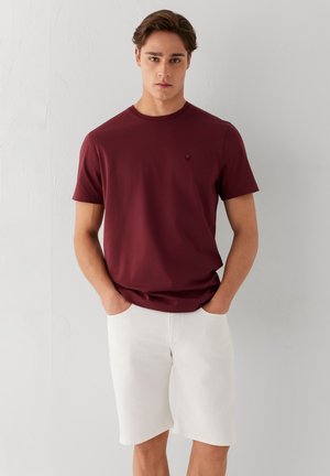 Junger Mann mit braunen Haaren, der ein dunkelrotes kurzärmeliges T-Shirt und weiße Shorts trägt und mit den Händen in den Taschen vor einer schlichten Wand steht.