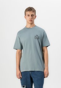 Fiatal férfi rövid hajjal, világoskék "Rip Curl Australia" pólót és szakadt kék farmert visel, fehér háttér előtt áll.