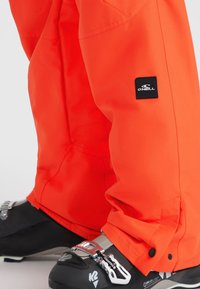 Felle oranje skibroek van waterafstotende stof, met een zwart logo patch en verstelbare manchetten, gecombineerd met zwarte skischanen.