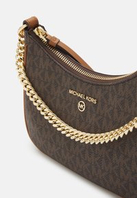 Ruskea kuvioitu käsilaukku, jossa on kultaketjuolkaimet, vetoketjusuljin ja tan-värinen nahkakoriste. Ominaisuuksiin kuuluu Michael Kors -brändäys kullassa.