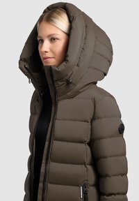 Pikowana parka w ciemnozielonym kolorze, z dużym, odpinanym kapturem, suwakiem z przodu oraz detalem z logo na rękawie. Gładki, teksturowany materiał.