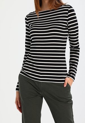Femme portant un t-shirt à manches longues noir et blanc à rayures horizontales avec un pantalon vert foncé, une main dans la poche et un bras détendu.