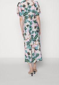 Mujer con un vestido de longitud media con mangas abullonadas y un patrón floral abstracto en verde, azul, rosa y marrón, combinado con sandalias de tacón beige.