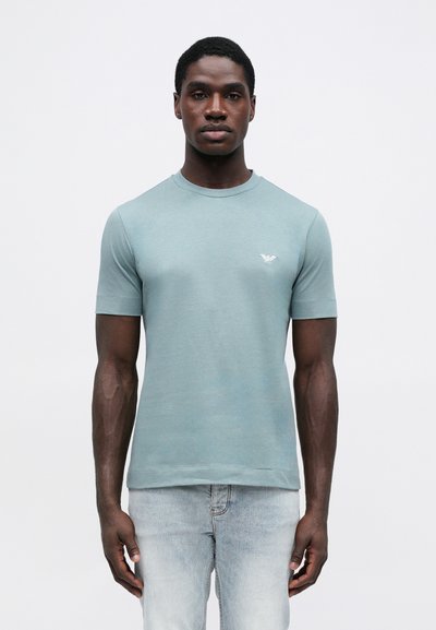 Svijetloplava majica kratkih rukava od mekog materijala, sa malim izvezenim logom na prsima. Dizajn s okruglim izrezom i prilagođen oblik.