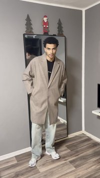 Manteau long beige clair à coupe décontractée, porté sur un t-shirt noir, associé à un pantalon en denim bleu clair et des baskets blanches.