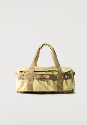 Geantă duffel galben deschis cu curele bej, fermoare laterale și închidere cu cataramă, așezată pe un fundal simplu gri deschis.