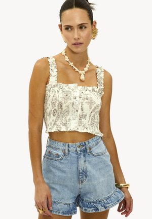 Femme portant un crop top floral crème avec des bretelles volantes, un short en denim taille haute bleu clair avec des ourlets volants, un collier en coquillage et des bijoux dorés.