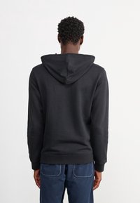 Svart hoodie med dragsko i huvan, mjukt tyg, långa ärmar, ribbade muddar och fåll. Bärs med blå byxor med synliga sömmar.