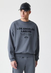 Šedá mikina s černým nápisem "LOS ANGELES LAKERS" na přední straně, s vroubkovanými manžetami a spodním lemem, doplněná o šedou čepici se stejným logem.