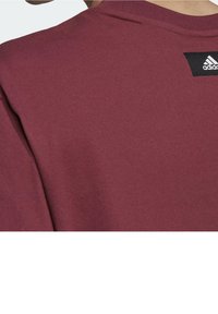 Mørkerød bomulds t-shirt med rund hals og korte ærmer, prydet med et sort Adidas-logo mærke på bagsiden nær halsudskæringen.