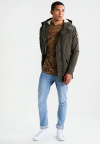 Giacca verde oliva con cappuccio foderato in pelliccia, t-shirt camouflage, jeans slim-fit azzurri chiari e sneakers bianche. Stile casual da esterno.