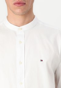 Man wearing a white buttoned shirt with a mandarin collar and a small red, white, and blue rectangular logo on the chest.