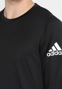 Mustapaita, jossa on pitkät hihat, pyöreä kaula-aukko, sileä rakenne ja valkoinen Adidas-logo vasemmassa hihassa.