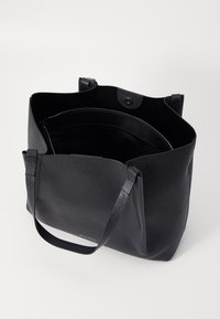 Sac fourre-tout en cuir noir avec texture granuleuse, ouverture en haut, deux poignées courtes et une poche zippée intérieure. Design minimaliste, sans logos visibles.