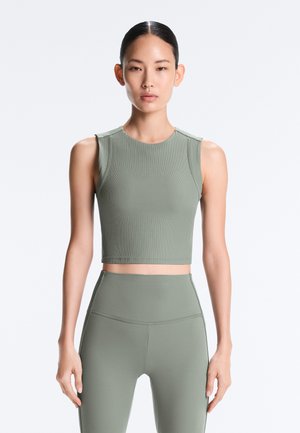 Woman stands facing forward wearing sleeveless ribbed crop top and high-waisted leggings in matching muted green on plain background.