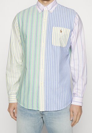 Chemise à manches longues boutonnée avec des rayures verticales vertes, jaunes, bleues et roses, portée avec un jean bleu clair.