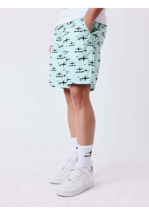 Uomo che indossa pantaloncini azzurri con motivi di stelle nere, calzini bianchi con logo nero e scarpe da ginnastica bianche, in piedi con le mani nelle tasche.