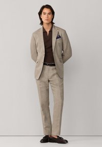 Muž v béžovom obleku, hnedej košeli, čiernom opasku, tmavomodrom vreckovke a tmavohnedých mokasínach s bruškami, stojaci pred neutrálnym pozadím.