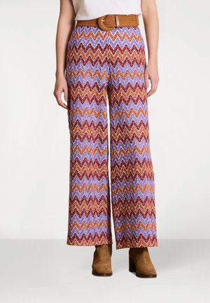 Pantalon à jambes larges à motifs, présentant un design en zigzag rouge, orange et bleu sur un fond clair ; complété par une ceinture tressée marron et des bottes marron.