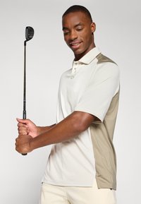 Polo de golf beige et marron clair à manches courtes, avec un col boutonné et des accents en panneaux latéraux. Figure masculine tenant un club de golf.