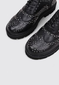 Schwarze Leder-Oxford-Schuhe mit silbernem Nietenakzenten, abgerundetem Zehenbereich, flacher Gummisohle und traditionellem Schnürdesign.