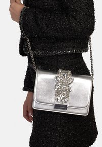 Srebrna metaliczna torebka na łańcuszkowym pasku, z ozdobn�ą, zdobioną klamrą i gładką fakturą, trzymana na tle czarnego, teksturowanego materiału.