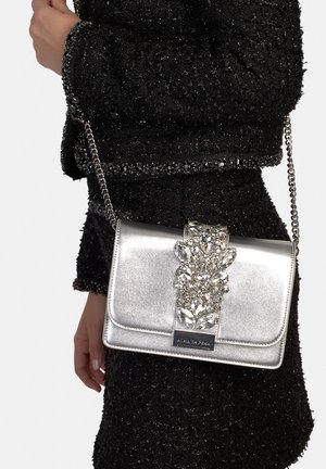 Borsa a tracolla in metallo argento con catenella, caratterizzata da una chiusura decorativa impreziosita e da una texture liscia, tenuta contro un tessuto nero texturizzato.