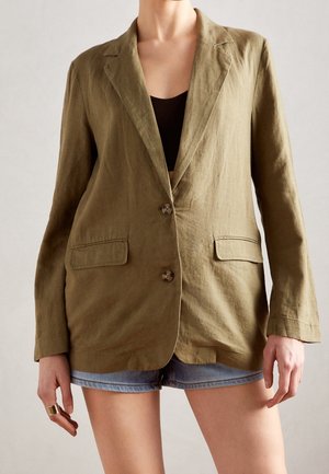 Olivgrüner Leinenblazer mit Reverskragen, zwei Vorderentaschen und zwei Knöpfen. Über einem schwarzen Top getragen und mit Jeansshorts kombiniert.