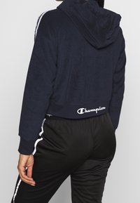 Felpa cropped blu navy in morbido tessuto con cappuccio, caratterizzata da strisce laterali bianche e un logo a contrasto all'orlo. Abbinata a pantaloni neri.