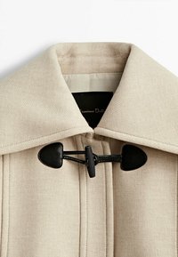 Beżowy płaszcz z mieszanki wełny z szerokim kołnierzem. Posiada czarne zapięcie na toggle oraz gładką teksturę z czystymi liniami. Podszewka widoczna wewnątrz.