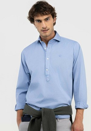 Hombre con cabello rizado y castaño lleva una camisa azul claro de manga larga con dos botones abiertos, un suéter gris atado alrededor de la cintura y pantalones de color claro.