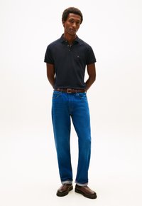 Tamsiai mėlyni polo marškinėliai, mėlyni džinsai, rudas diržas ir tamsūs batai. Modelis stovi susikabinęs rankas šviesiame fone.