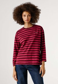 Bordo majica dugih rukava s vodoravnim ružičastim prugama, okruglim izrezom i malim logotipom na prsima. Nošena s tamnoplavim trapericama.