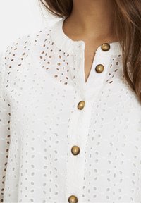 Weiße Bluse mit Lochstickerei, rundem Ausschnitt und braunen dekorativen Knöpfen entlang der Vorderseite. Leichtes und strukturiertes Material.