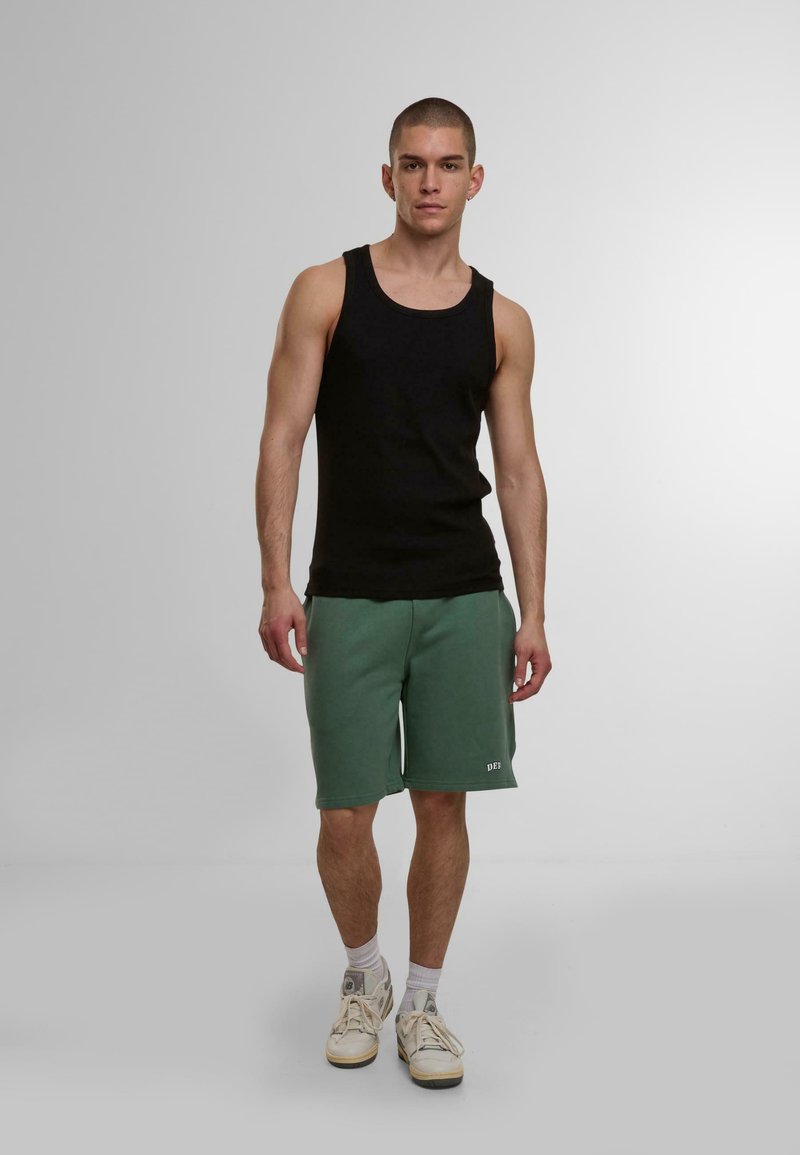 Schwarzes Tanktop kombiniert mit grünen Sweatshorts, die einen elastischen Bund und Seitentaschen haben. Als Schuhwerk werden hellfarbene Sneakers getragen.