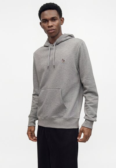 Jeune homme portant un sweat à capuche gris clair avec une poche avant et un petit logo zèbre sur la poitrine, debout devant un fond blanc uni.