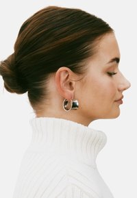 Sudraba auskari gredzenu formā ar spīguļojošu, atreflectējošu apdari, kas papildina baltu ribotu augsto apkakliņu. Mati sakārtojami zemu bunā.