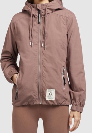 Vrouw met een mauve capuchonjack met trekkoorden, een rits aan de voorkant, ritszakken aan de zijkant en elastische manchetten, staand met één hand in de zak.