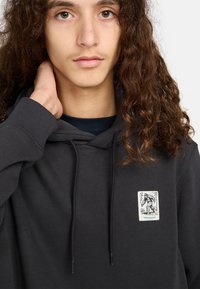 Jongere met lang krullend haar, gekleed in een zwarte hoodie met daarop een kleine afbeelding van Bigfoot en het woord "element."