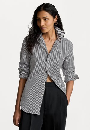 Mujer vistiendo una camisa de botones con rayas verticales en blanco y negro, con el cuello levantado y las mangas arremangadas, combinada con pantalones negros, con la mano en el bolsillo.