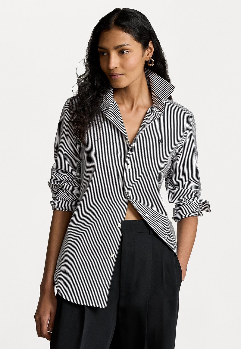 Femme portant une chemise à rayures verticales noir et blanc, avec le col relevé et les manches retroussées, assortie à un pantalon noir, la main dans la poche.