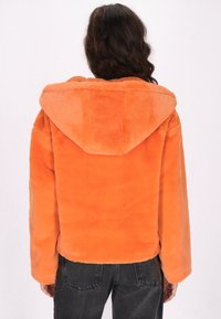 Giacca con cappuccio in pelliccia sintetica arancione, con una superficie morbida e testurizzata e maniche lunghe, caratterizzata da un orlo arrotondato, indossata sopra jeans scuri.