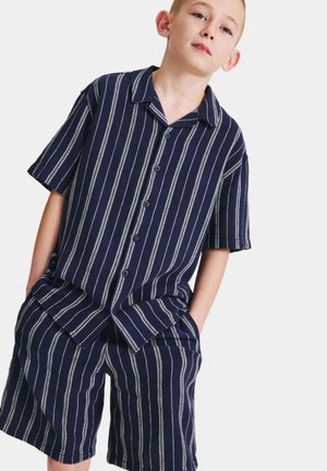 Junge trägt ein marineblaues, kurzärmeliges Hemd mit Knöpfen und passende Shorts mit weißen vertikalen Streifen, Hände in den Taschen vor weißem Hintergrund.