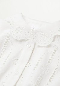 Blouse en coton blanc avec col en broderie anglaise festonné, et panneaux en dentelle ajourée verticale, dotée de boutons sur le devant.