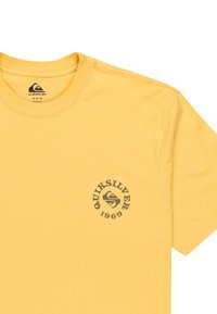 Žluté bavlněné tričko s kulatým výstřihem. Na levé části hrudi se nachází kruhové logo Quiksilver v tmavě šedé barvě. Design s klasickým střihem.